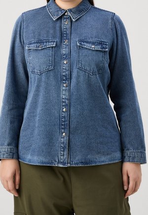Frau trägt ein blaues Jeanshemd mit Knöpfen und zwei Brusttaschen und olivgrüne Hose, steht vor einem einfarbigen Hintergrund.