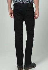 Muž v černých slim-fit džínách a černých kožených botách, stojící zády k světlému šedému pozadí.