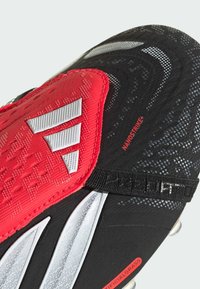 Μαύρο ποδοσφαιρικό παπούτσι με κόκκινες και λευκές λεπτομέρειες. Διαθέτει υφασμένο επάνω μέρος, τρεις λευκές ρίγες και έναν ιμάντα με την ετικέτα "PREDATOR".