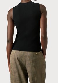 En person står klädd i en svart ribbad ärmlös topp och ljusa rutig byxor, vilket visar bakvyn av outfiten mot en neutral bakgrund.