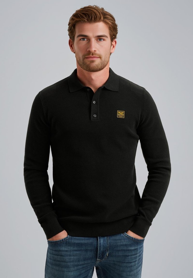 Zwarte lange mouwen polo met een gestructureerde brei, drie knoopsluitingen en een gele labelpatch op de borst, gecombineerd met blauwe jeans.