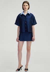 Navy satin short-sleeve shirt with light blue collar, two front pockets, paired with a matching short skirt and black heeled sandals.