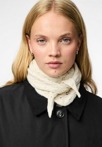 Uldscarf i creme, viklet omkring halsen med et strikket mønster, båret over en sort frakke med knaplukning og krave.