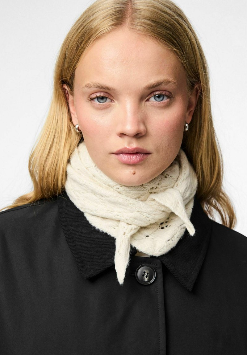 Uldscarf i creme, viklet omkring halsen med et strikket mønster, båret over en sort frakke med knaplukning og krave.
