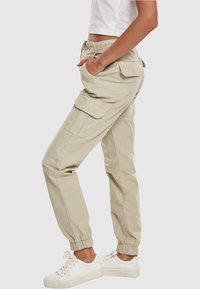 Boční pohled na osobu v béžových cargo kalhotách s elastickými manžetami a bílými teniskami, ruka v kapse, na jednobarevném pozadí.