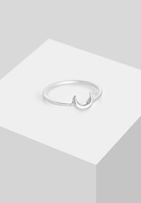 Bague en argent avec un design de croissant de lune reposant sur un piédestal géométrique blanc sur un fond gris clair.