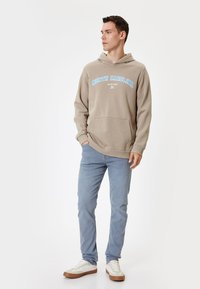 Sudadera beige con el texto "NORTH CAROLINA" en azul claro, bolsillo frontal, combinada con jeans azul claro y zapatillas blancas con suela marrón.