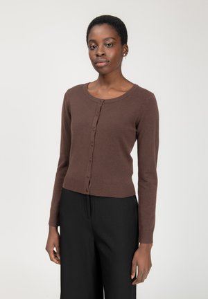 Cardigan en tricot marron avec manches longues, encolure ronde et fermeture par boutons. Présente un ourlet et des poignets côtelés. Porté avec un pantalon noir.