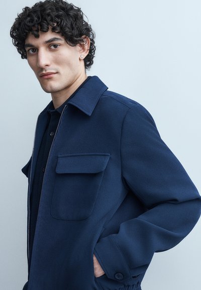 Νέος άνδρας με σγουρά σκούρα μαλλιά που φοράει μπλε σκούρο μπουφάν με μπροστινή τσέπη και μανσέτες με κουμπιά, στέκεται μπροστά σε ανοιχτό γκρι φόντο.