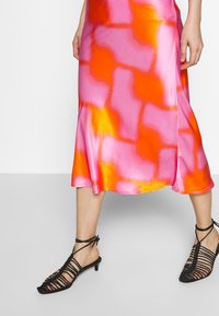 Jupe midi en satin avec un motif abstrait rose et orange, associée à des sandales à talons ouvert noires avec des lanières.
