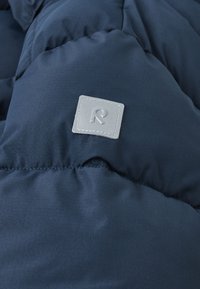 Tessuto imbottito blu navy con texture trapuntata, caratterizzato da una toppa quadrata grigia con un logo "R" stilizzato. Materiale morbido e liscio.