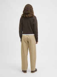 Mulher com cabelo encaracolado até aos ombros veste um suéter castanho, calças largas bege e sapatos castanho-escuros, em pé contra um fundo branco liso.