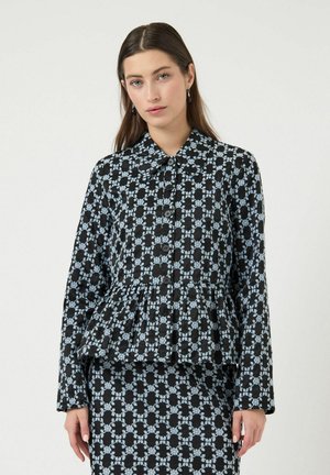 Mujer joven vestida con un conjunto negro con un patrón geométrico floral azul claro, mangas largas, cuello y botones al frente, de pie contra un fondo blanco.