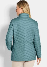Woman with blonde hair wearing a teal quilted jacket viewed from the back, standing against a light gray background.