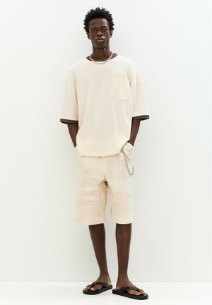 Light beige textured t-shirt with short sleeves and a chest pocket, paired with beige shorts and black flip-flops. Silver chain accessory.