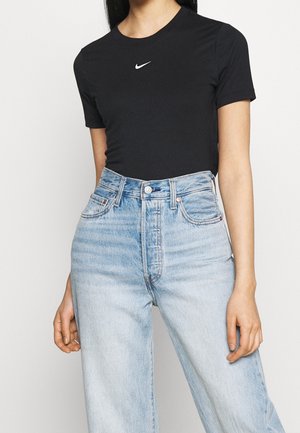 Frau trägt schwarzes Nike-T-Shirt mit kurzen Ärmeln, in hoch taillierte hellblaue Jeans gesteckt, steht vor einfarbigem Hintergrund.