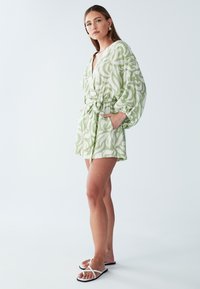 Groene romper met patroon, een V-hals, cinched taille en brede mouwen. Gelaagde stof met zakken, gedragen met witte flip-flops.