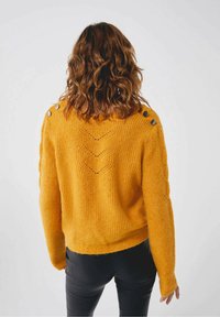 Femme aux cheveux bruns ondulés portant un pull tricoté jaune moutarde avec des boutons sur les épaules et un pantalon noir, vue de dos.