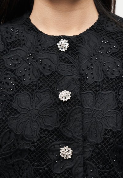 Μαύρο υφασμάτινο ύφασμα με υφή και μικρές μαύρες χάντρες και τρία διακοσμητικά κουμπιά σε σχήμα κρυστάλλινων λουλουδιών σε ένδυμα.