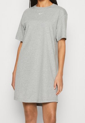 Žena v krátkém šedém šatu značky Nike s krátkými rukávy a délce po kolena, s malým bílým logem na hrudi, stojí před jednobarevným pozadím.