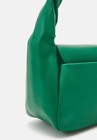 Zelená hladká kožená kabelka s krátkým, zkrouceným držadlem a viditelnými bočními švy, vyfotografována na bílém pozadí.