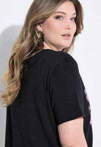 Černé tričko s krátkým rukávem a kulatým výstřihem. Na přední straně se nachází růžový květinový potisk a textura látky je jemná a hladká.