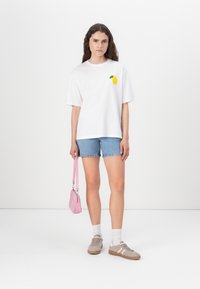 Camiseta blanca de manga corta con un gráfico de limón amarillo, combinada con pantalones cortos de denim, un bolso rosa y zapatillas beige con acentos rosas.