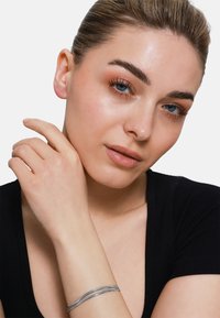 Silberne Armbanduhr mit einem glatten, gedrehten Design. Das Modell trägt ein enges schwarzes Top, das das Armband an ihrem Handgelenk zur Geltung bringt. Helle Haut und dezentes Make-up.