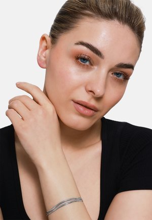 Bracciale d'argento con un design liscio e attorcigliato. Il modello indossa un top nero aderente, mettendo in mostra il bracciale sul suo polso. Carnagione chiara e trucco delicato.