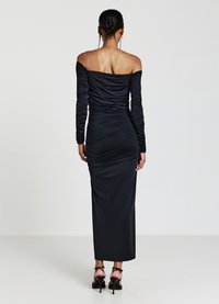Abito nero off-the-shoulder realizzato in tessuto morbido con dettagli arricciati. Il vestito è lungo fino al pavimento e abbinato a sandali con tacchi a strappo.