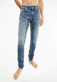 Světle modré džíny s úzkým střihem, které mají klasický pětikapsový design, se světlejšími oblastmi a zapínáním na knoflík.