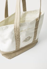 Lærret tote bag i kremfarge med en beige bunn. Har doble topphåndtak og en frontlomme med trykt logo. Sømdetaljer på sømmene.