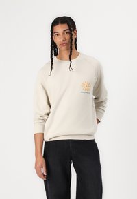 Helles BEIGE Sweatshirt mit einem gerippten Rundhalsausschnitt, Raglanärmeln und einem orange-blauen Sonnenmotiv auf der linken Brust. Texturiertes Material.