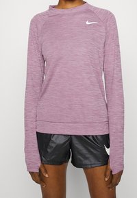 Langärmeliges, meliertes hellviolettes Sportshirt mit Rundhalsausschnitt, schmaler Passform und weißem Nike-Logo. Kombiniert mit schwarzen Sportshorts.