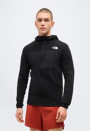 Giovane uomo con indosso una felpa nera con zip North Face e pantaloncini rossi, in piedi davanti a uno sfondo grigio chiaro uniforme.