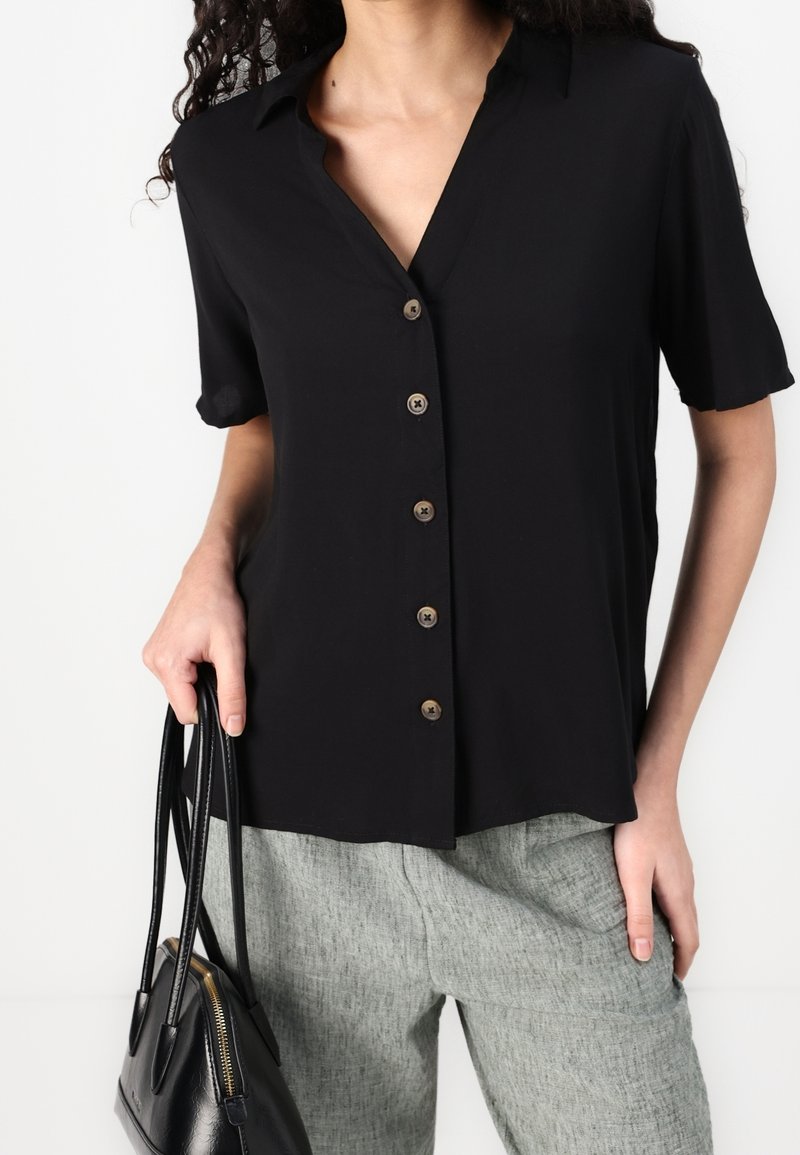 Femme portant une chemise noire à manches courtes boutonnée et un pantalon gris, tenant un sac à main noir dans sa main gauche.