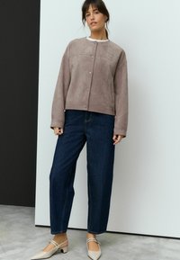 Taupe suède jas met een ronde halslijn en drukknoopjes, gecombineerd met donkerblauwe denim jeans en beige lage hakken.