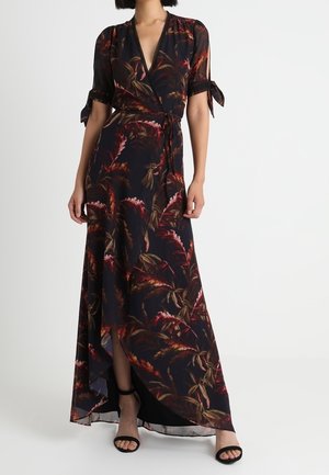 Mujer con un vestido envolvente floral oscuro de manga corta atada en los codos y tacones abiertos negros.
