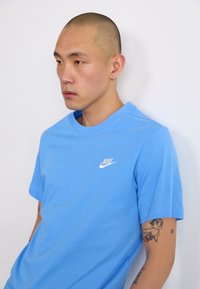 Modra kratkotrajna majica z belim logotipom Nike na levem prsnem delu, izdelana iz mehkega materiala. Vidna koža z tatuji na rokah.