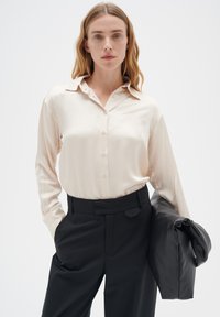 Hellbeige Hemd mit Knöpfen, Kragen und langen Ärmeln, kombiniert mit maßgeschneiderten schwarzen Hosen und einer schwarzen Jacke über dem Arm.