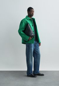 Zelena jakna iz volnene mešanice s koničastim ovratnikom, črna torbica, črna srajca, ohlapne modre jeans hlače in črne čevlje na sivi ozadju.