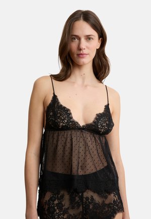 Donna che indossa lingerie nera in pizzo floreale e tulle a pois con spalline sottili, in piedi contro uno sfondo bianco.