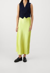 Gilet sans manches bleu marine avec une fermeture à bouton sur une longue robe en satin jaune vif. Des mocassins noirs complètent l'ensemble.
