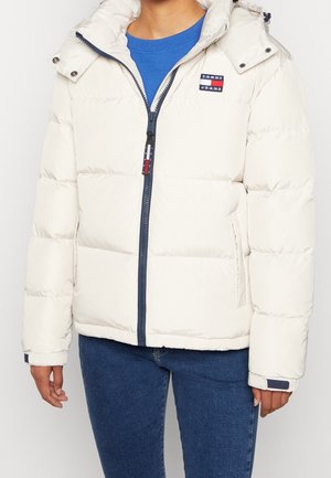 Persoon die een witte donsjas met capuchon en een marineblauwe rits draagt, over een blauw shirt en blauwe jeans, staand tegen een effen achtergrond.