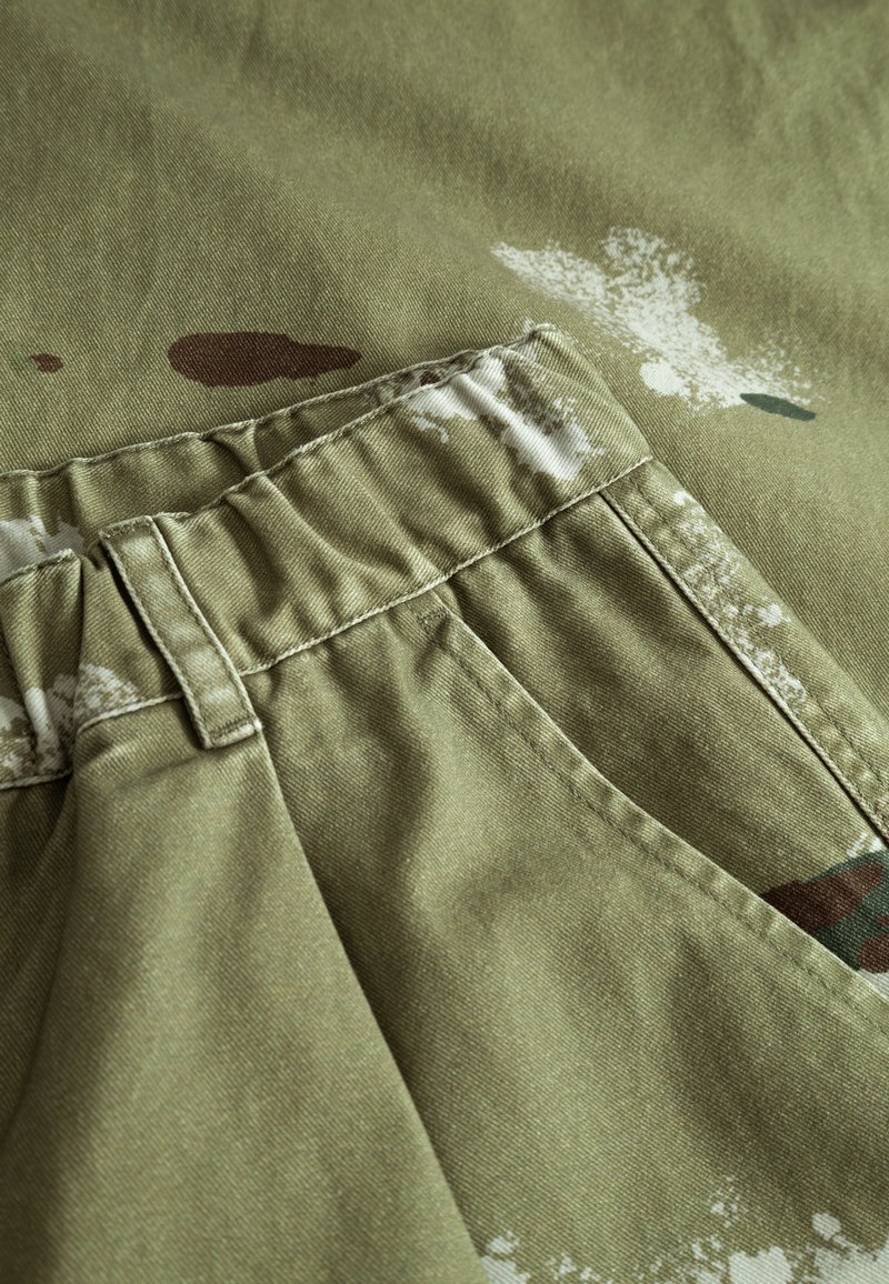 Khaki-stof med varierende nuancer og strukturerede mønstre, der har en samlet talje, sidelomme og abstrakte sprøjtedetaljer.