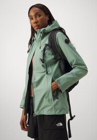 Veste imperméable vert clair avec capuche ajustable, fermeture éclair avant et poches latérales, portée avec un sac à dos noir et un short foncé.