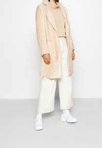 Abrigo de pelo sintético de un beige claro con cuello muesca, dos bolsillos frontales, llevado sobre un suéter de cuello alto beige y pantalones de pierna ancha blancos; combinado con zapatillas blancas.