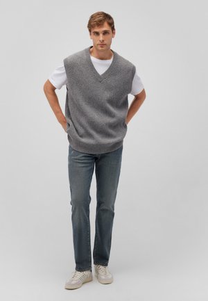 Grauer strickloser Pullover mit V-Ausschnitt, getragen über einem weißen T-Shirt, kombiniert mit blauen Jeans und weißen Sneakern, vor einem hellen Hintergrund stehend.