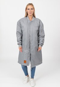 Vrouw staat in een lange lichtgrijze bomberjack, blauwe jeans en witte sneakers tegen een witte achtergrond.