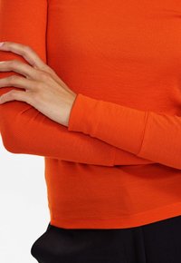 Personne aux bras croisés portant un haut à manches longues côtelé de couleur orange vif et un pantalon noir, sur un fond blanc uni.